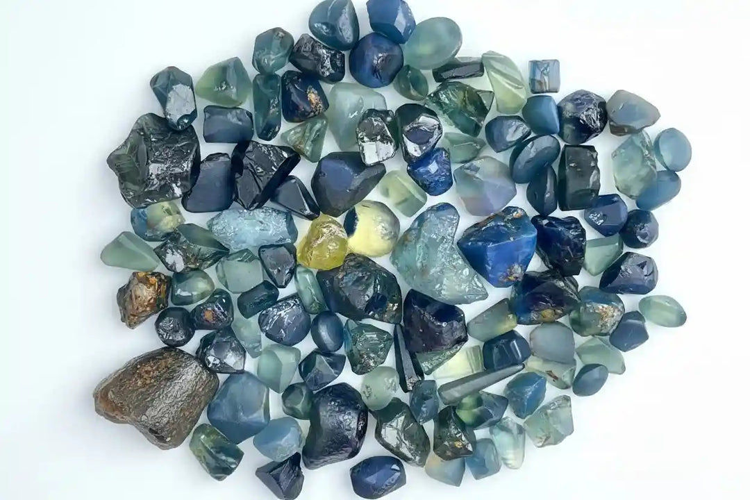 A collection of natural rough teal and blue sapphire gemstones arranged on a white background