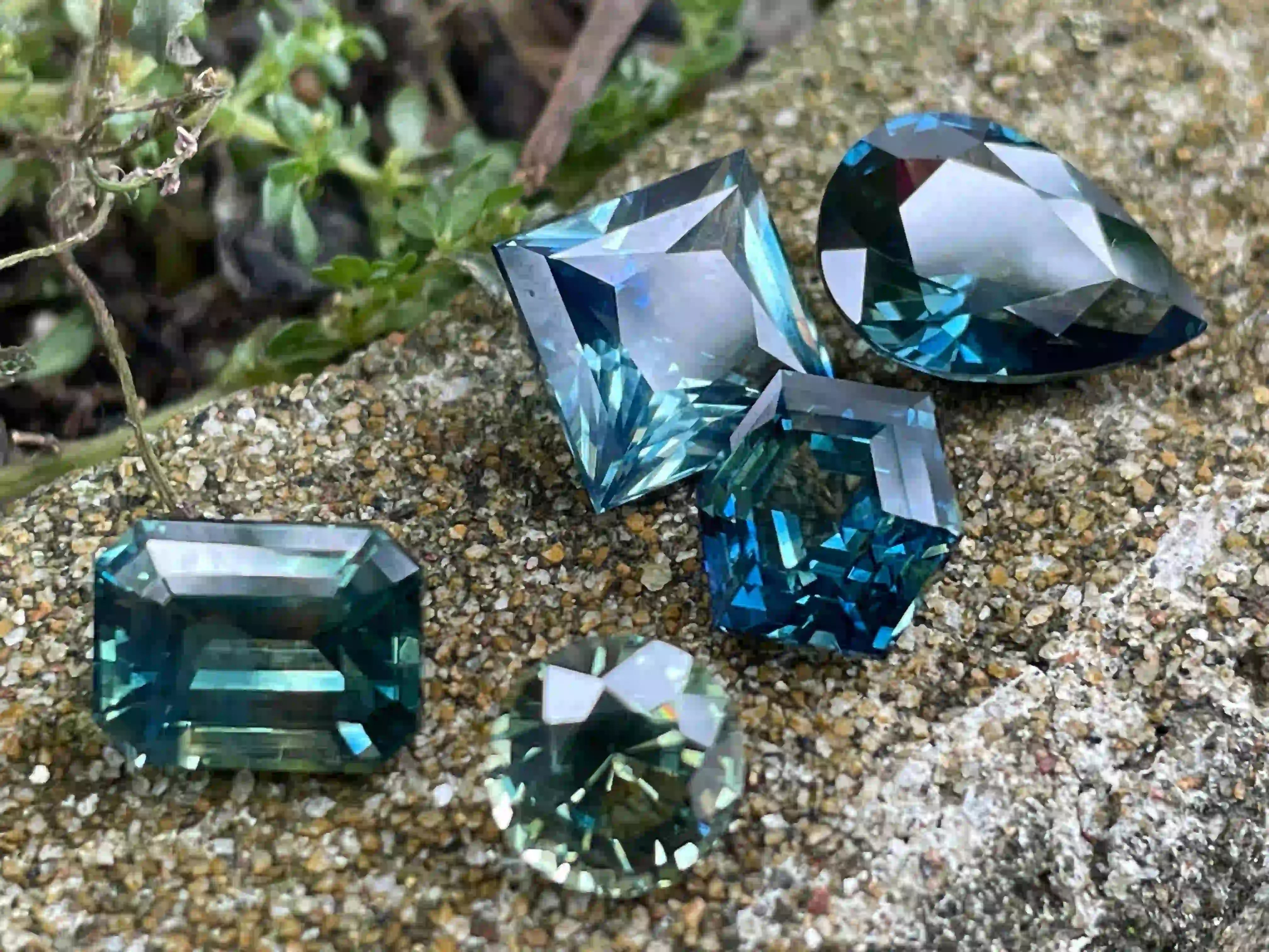 A collection of teal and bi-color parti sapphires in various cuts, including emerald, pear, and hexagon, displayed on a textured stone surface in natural daylight